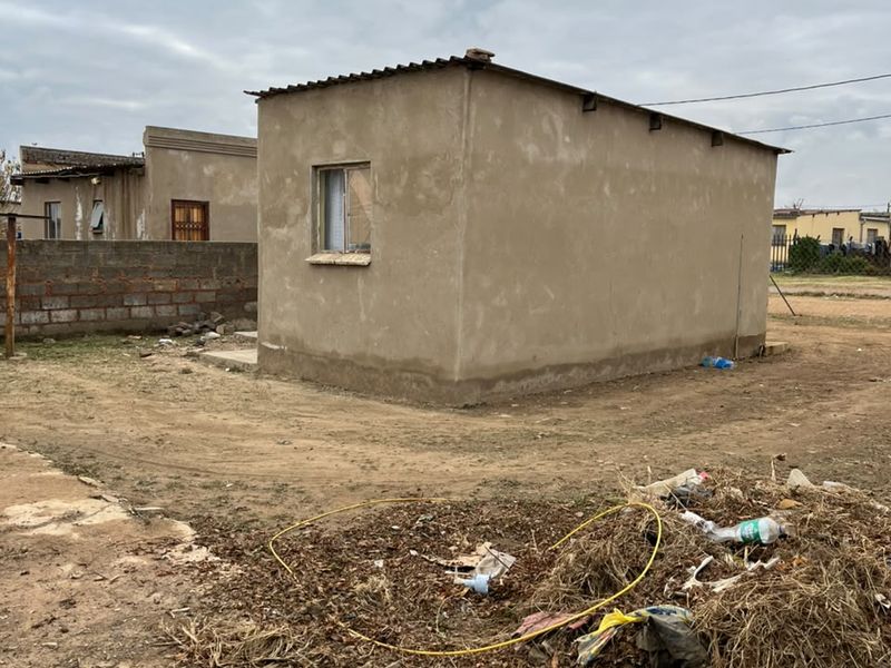 A detached structure with cement rendered walls and a corrugated metal roof, situated on an unpaved lot with visible debris.