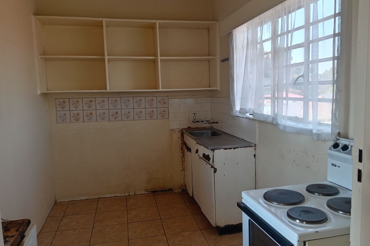 A dated kitchen featuring basic cabinetry, a stainless steel sink, open shelving, and tiled flooring, requiring renovation to modernize the space.