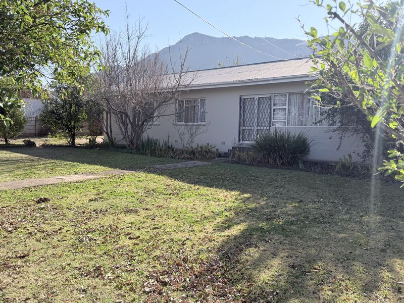Rear view of a single-story house featuring a metal roof, security gate, and established garden with mountain views.