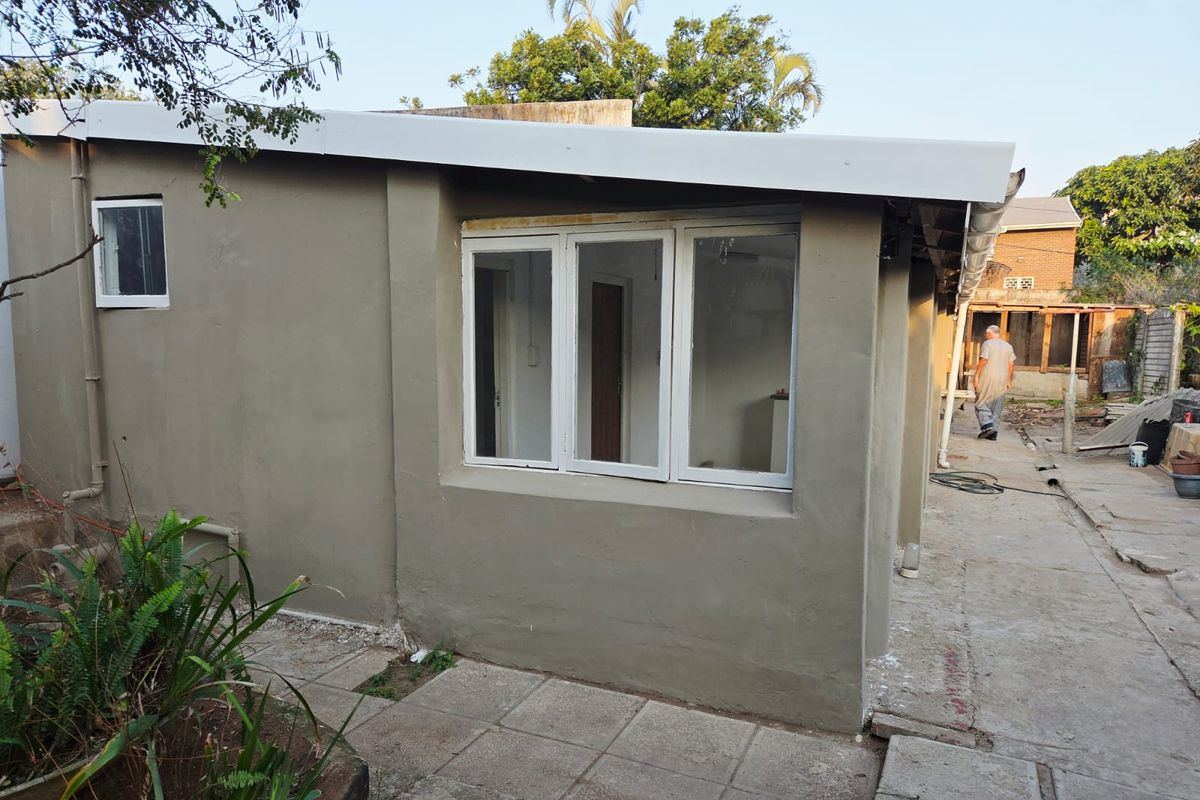 Rear exterior view of the property showing plastered walls, aluminium window frames, and concrete paving in a state of ongoing maintenance or renovation.