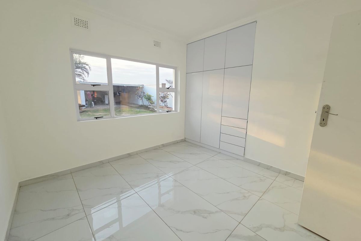 A bright, empty bedroom featuring modern white marble-look floor tiles, built-in cupboards, and large aluminium-framed windows overlooking the rear garden.
