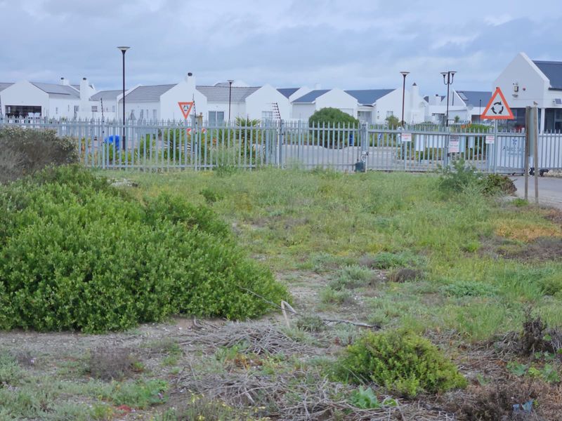 A view of a gated residential estate entrance featuring metal security fencing and street lighting, surrounded by natural coastal vegetation.
