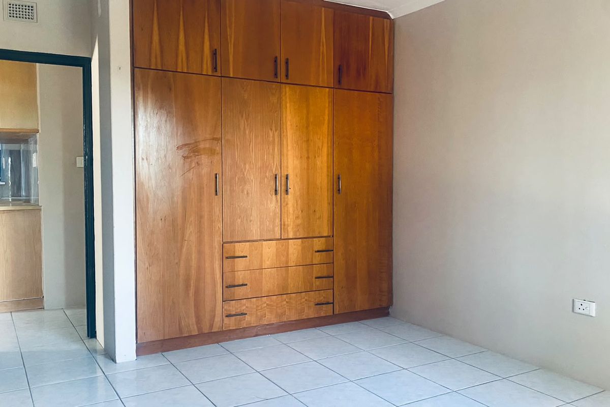 A bedroom featuring built-in wooden wardrobes and tiled flooring, with a view into the adjacent kitchen area.
