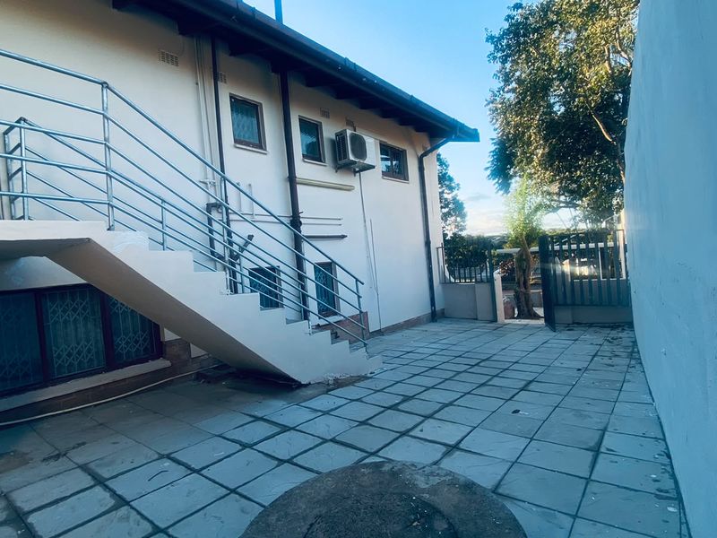 Rear exterior view of the cottage featuring a paved patio area, external staircase with metal railings, and wall-mounted air conditioning unit.