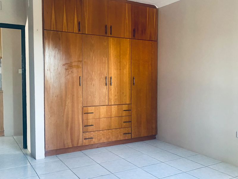 A bedroom featuring built-in wooden wardrobes and tiled flooring, with a view into the adjacent kitchen area.