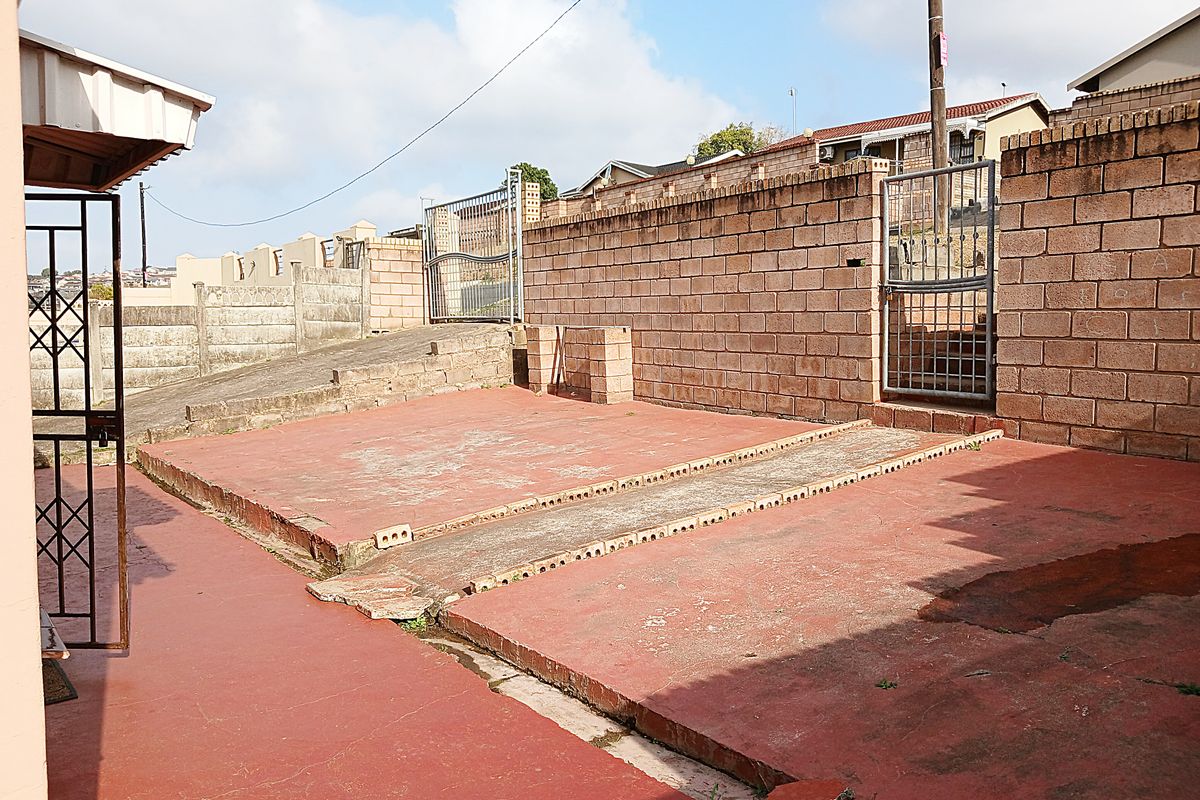 Rear exterior area featuring a concrete driveway, brick boundary walls, and metal security gates, showing signs of wear and surface weathering.
