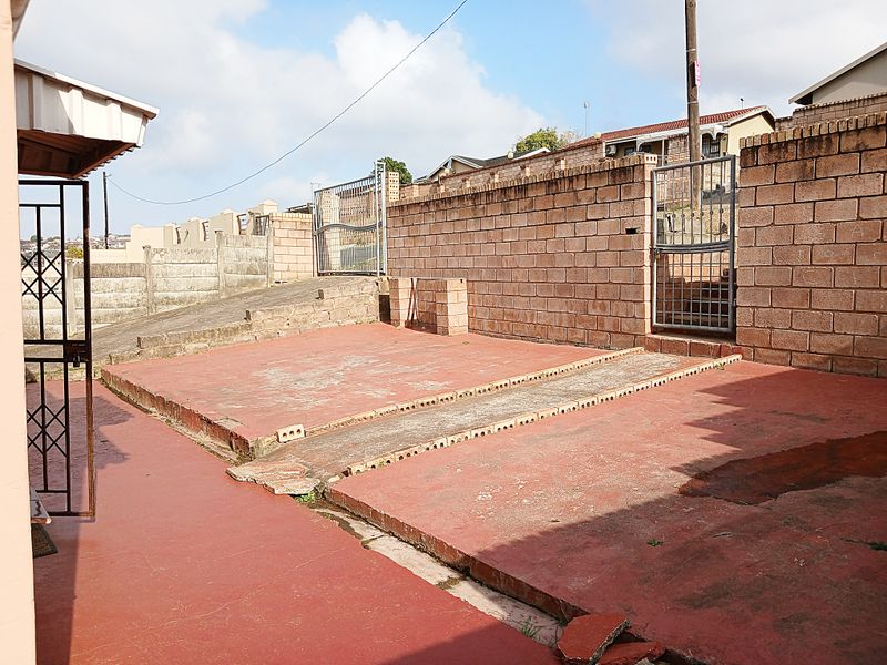 Rear exterior area featuring a concrete driveway, brick boundary walls, and metal security gates, showing signs of wear and surface weathering.