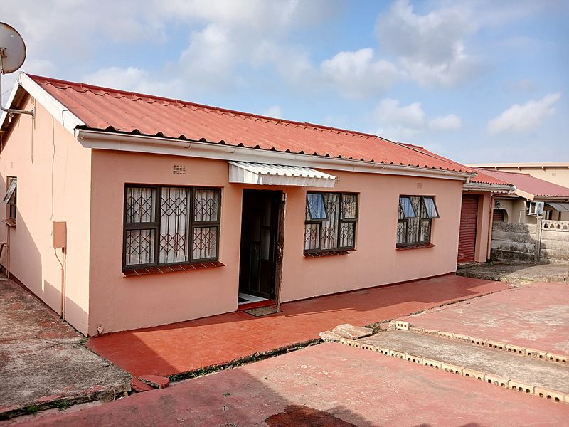 Front exterior view of a single-story house featuring a red corrugated metal roof, security-barred windows, and a paved driveway area.