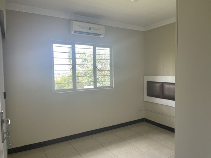 A clean, empty bedroom featuring tiled floors, a wall-mounted air conditioning unit, and a built-in headboard with security bars on the window.