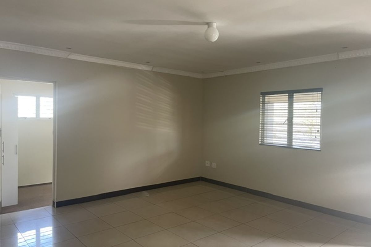 A spacious, empty living area featuring neutral tiled flooring, crown molding, and natural light through a window with fitted blinds.