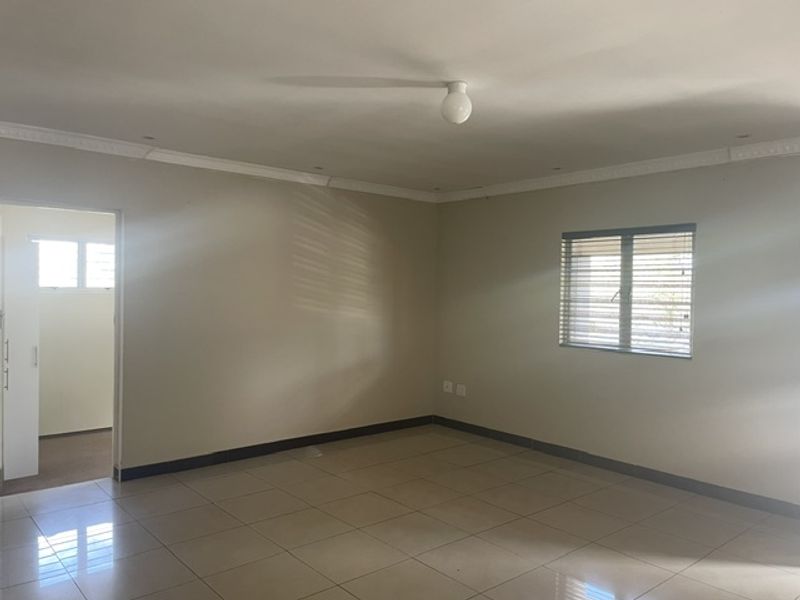 A spacious, empty living area featuring neutral tiled flooring, crown molding, and natural light through a window with fitted blinds.