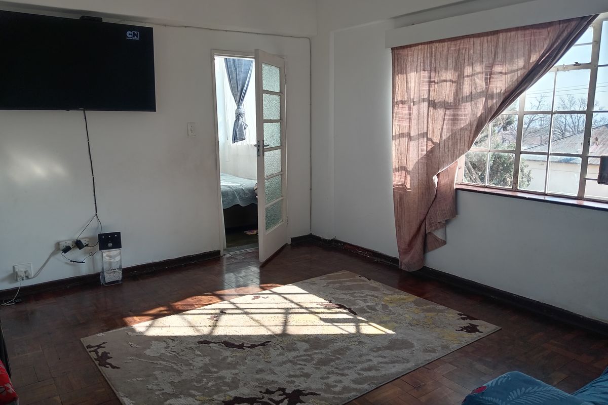 A living room featuring original parquet flooring, large steel-framed windows, and direct access to an adjoining room.