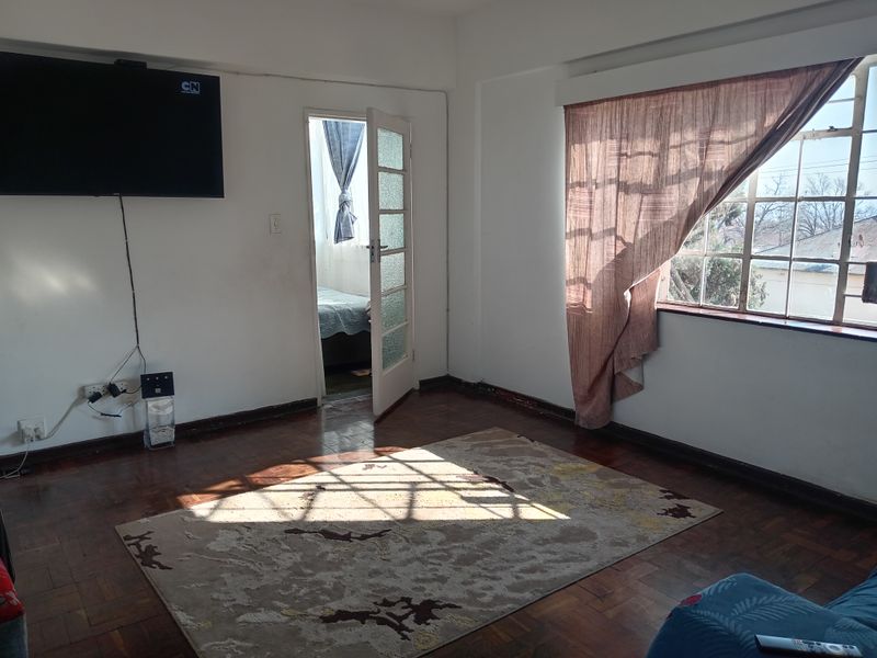 A living room featuring original parquet flooring, large steel-framed windows, and direct access to an adjoining room.