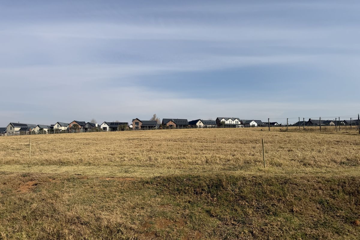 A wide-angle view of a vacant residential plot featuring dry grassland with established residential homes visible in the background.