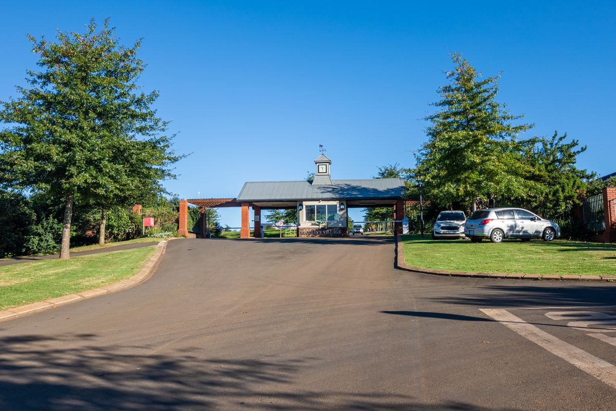 The main entrance and security gatehouse of St John's Village, featuring a well-maintained asphalt driveway, landscaped grounds, and a prominent clock tower structure.