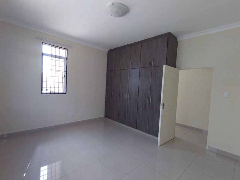 A bright, empty bedroom featuring neutral tiled flooring, modern dark wood built-in cupboards, and a window fitted with security bars.