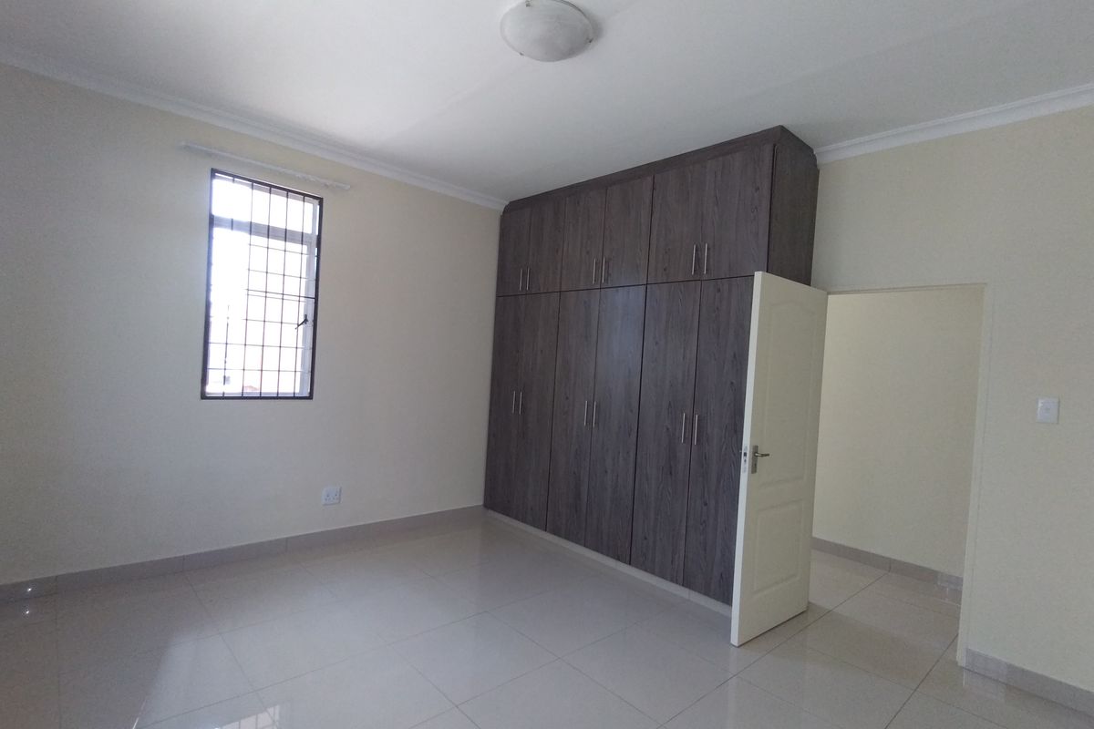 A clean, empty bedroom featuring modern dark wood built-in cupboards, neutral tiled flooring, and a window with security bars.