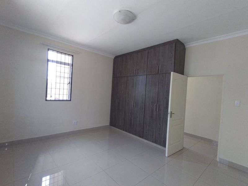 A clean, empty bedroom featuring modern dark wood built-in cupboards, neutral tiled flooring, and a window with security bars.