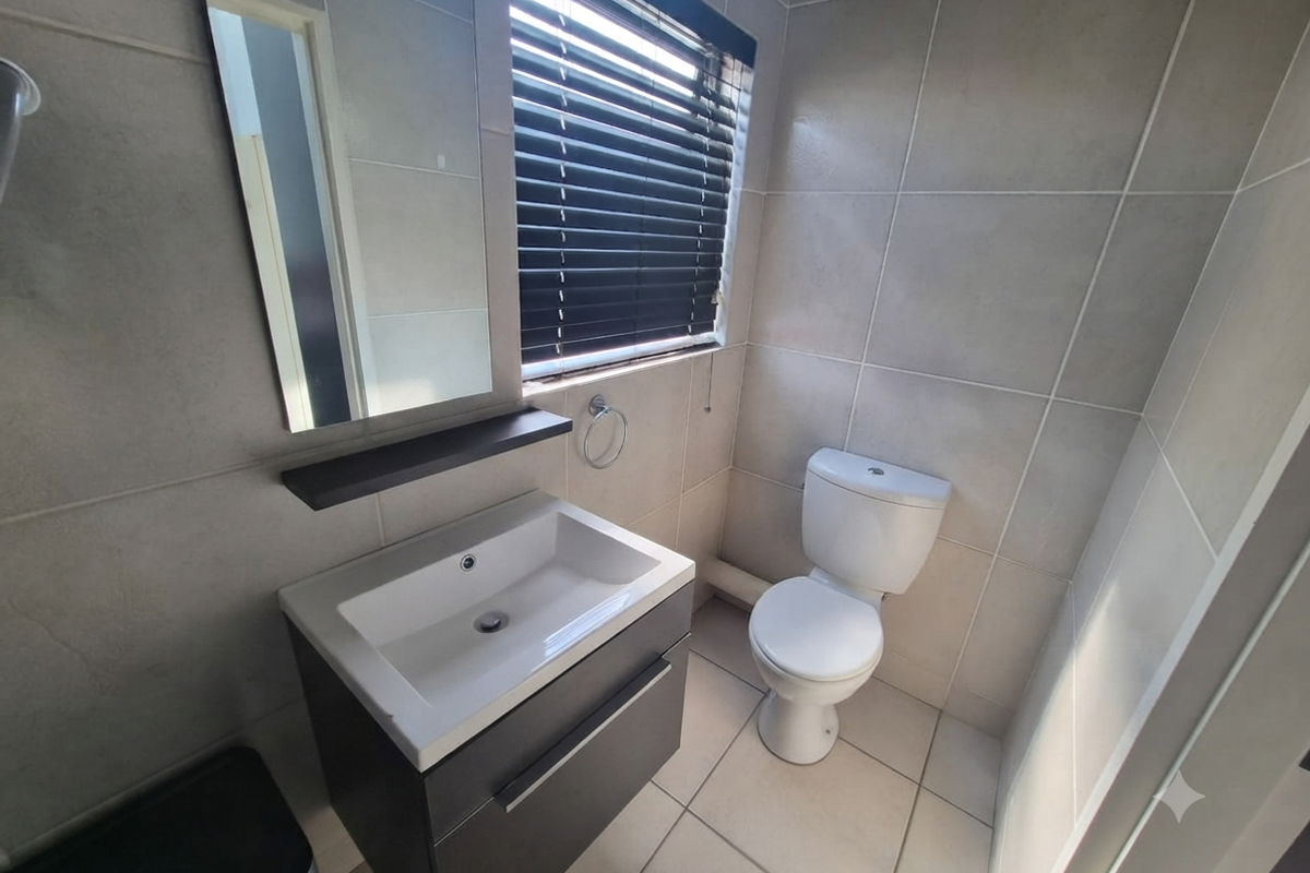 A clean, modern bathroom featuring a vanity cabinet with a basin, wall-mounted mirror, toilet, and neutral floor-to-ceiling tiling.