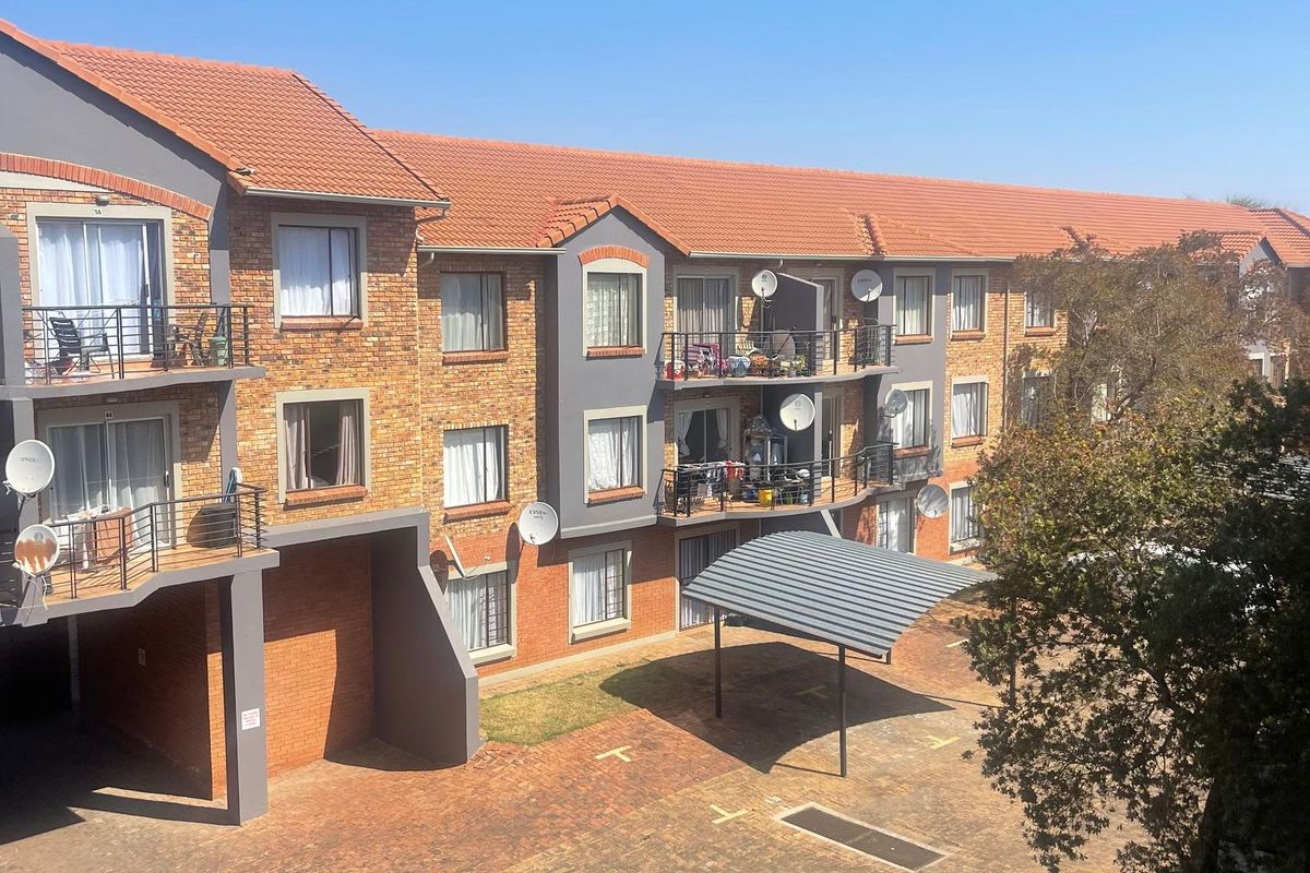 A view of the apartment complex exterior featuring brick facades, tiled roofing, private balconies with metal railings, and a paved parking area with carports.