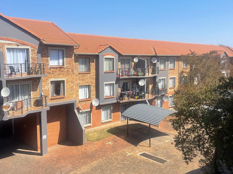 A view of the apartment complex exterior featuring brick facades, tiled roofing, private balconies with metal railings, and a paved parking area with carports.