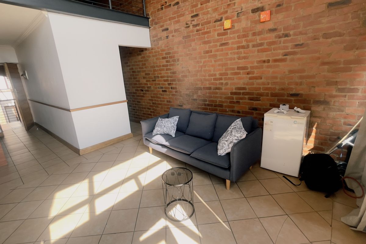 Spacious living area featuring an industrial-style exposed brick wall, tiled flooring, and a mezzanine level with a metal railing.