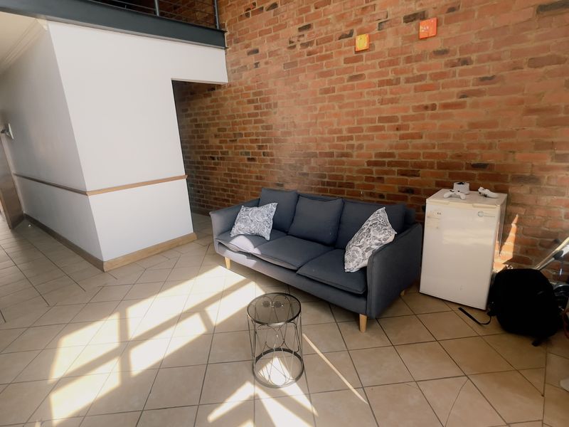Spacious living area featuring an industrial-style exposed brick wall, tiled flooring, and a mezzanine level with a metal railing.