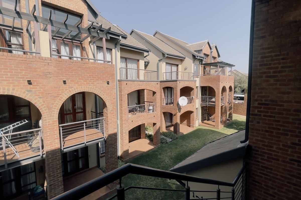 A view of the rear apartment complex facade featuring exposed brickwork, arched architectural details, private balconies with metal railings, and a well-maintained communal garden area.