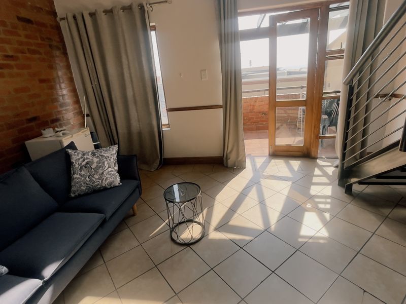 Living room featuring an exposed brick wall, tiled flooring, and a staircase with metal railings, leading out to a balcony through a glass door.