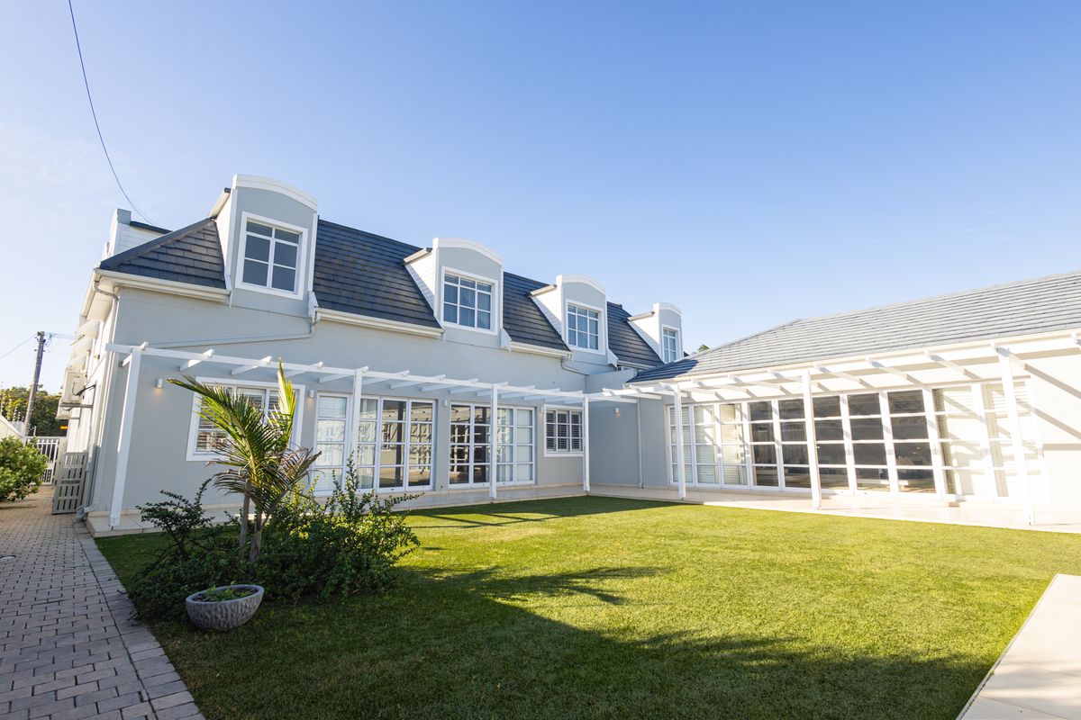 A well-maintained rear exterior view featuring a spacious lawn, paved pathways, and a white pergola structure attached to the main house with french doors.