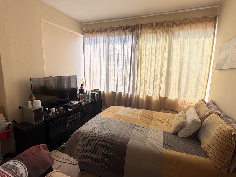 A bedroom featuring tiled flooring and a large window, currently showing signs of wear on the walls and plaster.