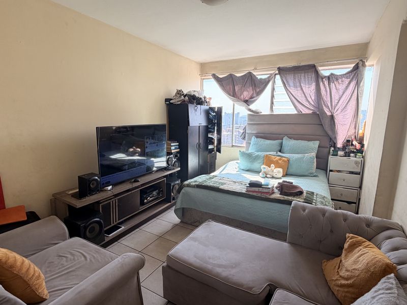 A bedroom featuring tiled flooring and a large window, currently used as a multi-functional living and sleeping space.
