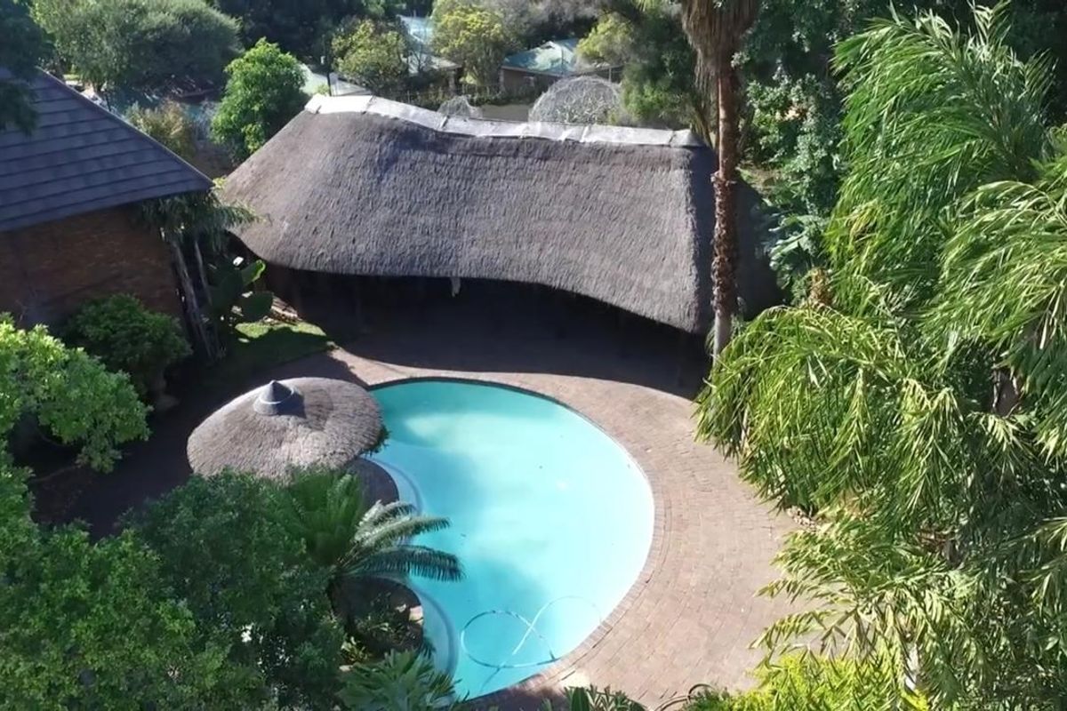 Aerial view of a swimming pool area featuring a large thatched lapa, paved surround, and established garden setting.