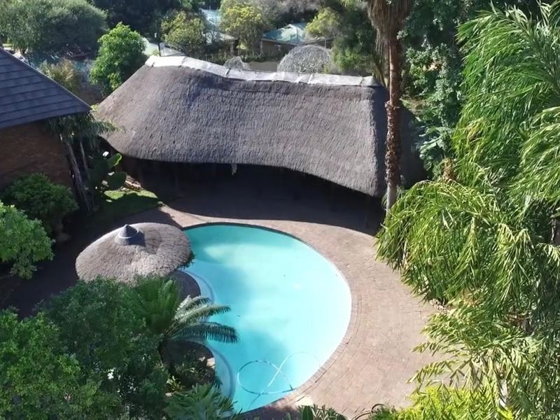 Aerial view of a swimming pool area featuring a large thatched lapa, paved surround, and established garden setting.