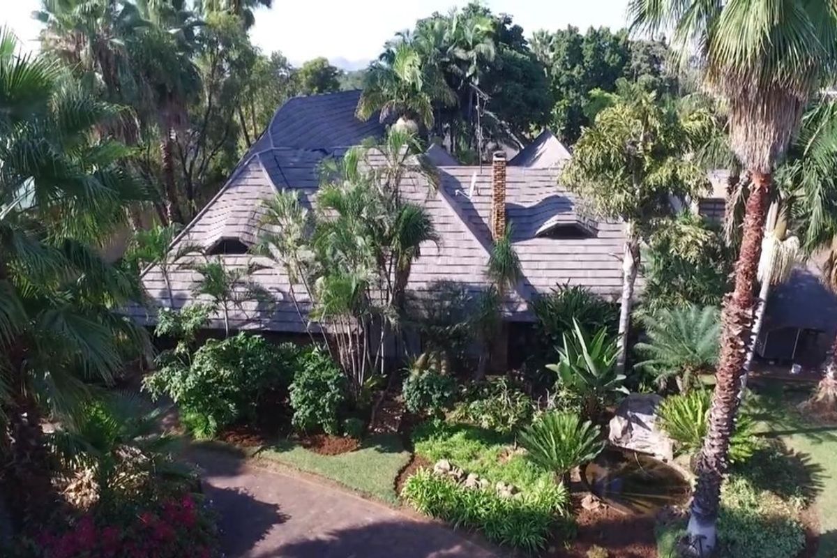 Aerial view of a residential property featuring a tiled roof, brick chimney, dormer windows, and a lush, landscaped garden with a paved driveway.