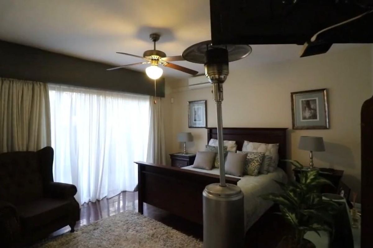 A spacious bedroom featuring hardwood flooring, a ceiling fan, and a wall-mounted split-system air conditioner, with large windows dressed in curtains.