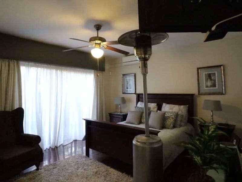 A spacious bedroom featuring hardwood flooring, a ceiling fan, and a wall-mounted split-system air conditioner, with large windows dressed in curtains.
