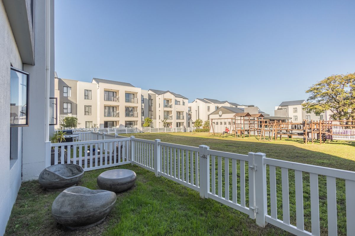 A view of a well-maintained communal garden area featuring a lawn, children's playground, and white picket fencing within an apartment complex.