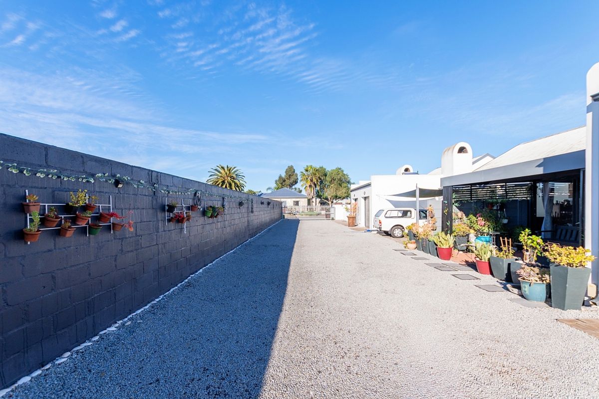 A spacious rear exterior area featuring a long gravel driveway, a high boundary wall, and a covered patio space.