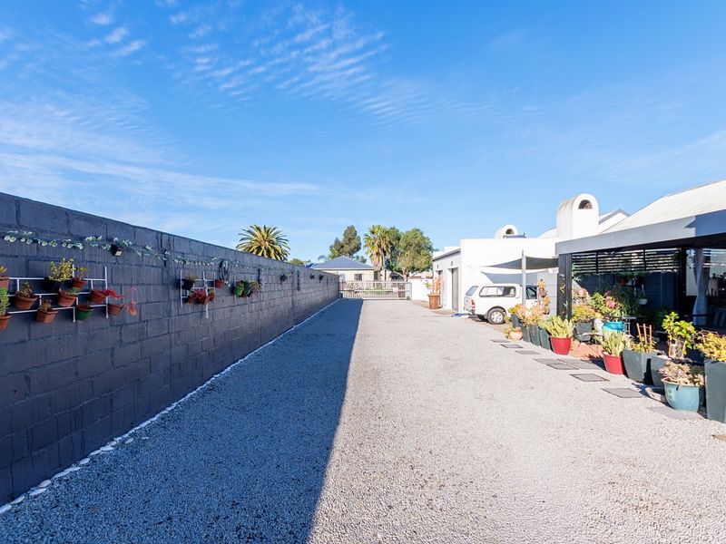 A spacious rear exterior area featuring a long gravel driveway, a high boundary wall, and a covered patio space.
