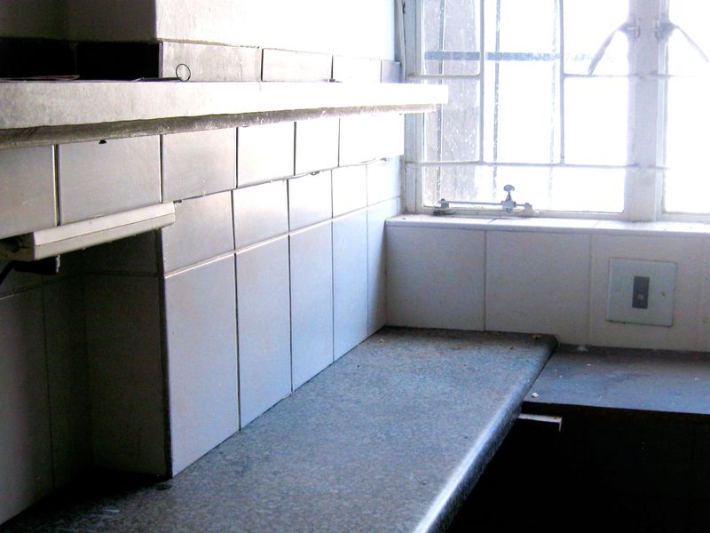 A dated kitchen corner featuring a tiled splashback, laminate countertop, and a steel-framed window, requiring cosmetic updates to modernize the space.
