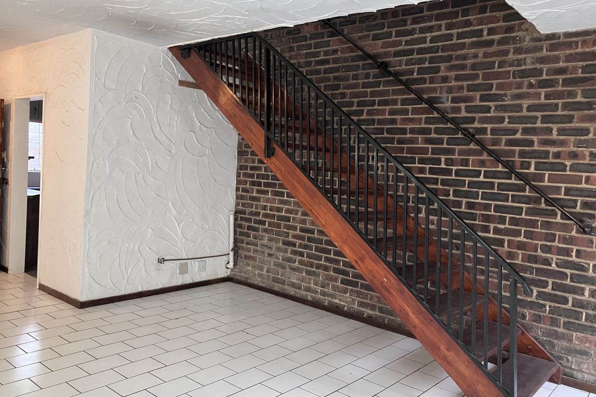 An open-plan living area featuring an exposed brick wall, a wooden staircase with a metal handrail, and tiled flooring.