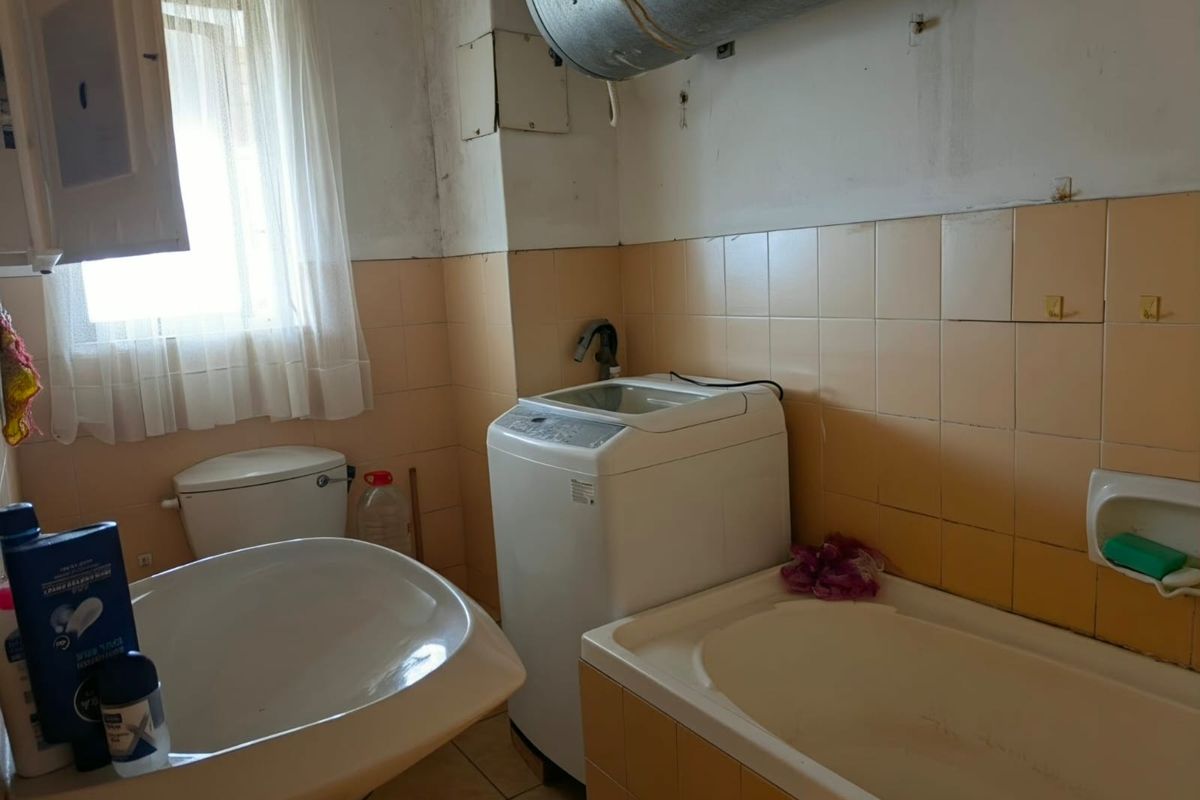 A dated bathroom featuring a bathtub, toilet, and ceramic tiling, currently showing signs of wear and exposed utility infrastructure.