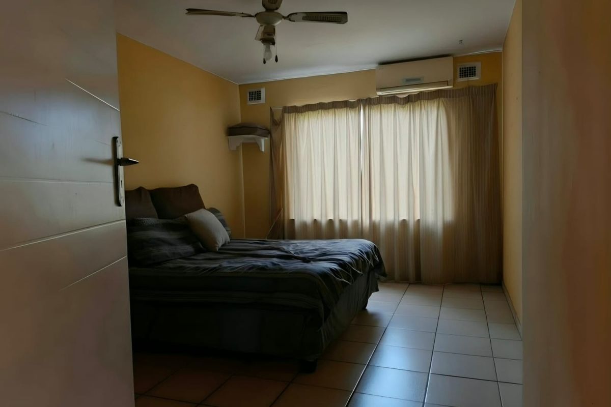 A bedroom featuring tiled flooring, a ceiling fan, and a wall-mounted air conditioning unit, providing a functional space with neutral wall finishes.