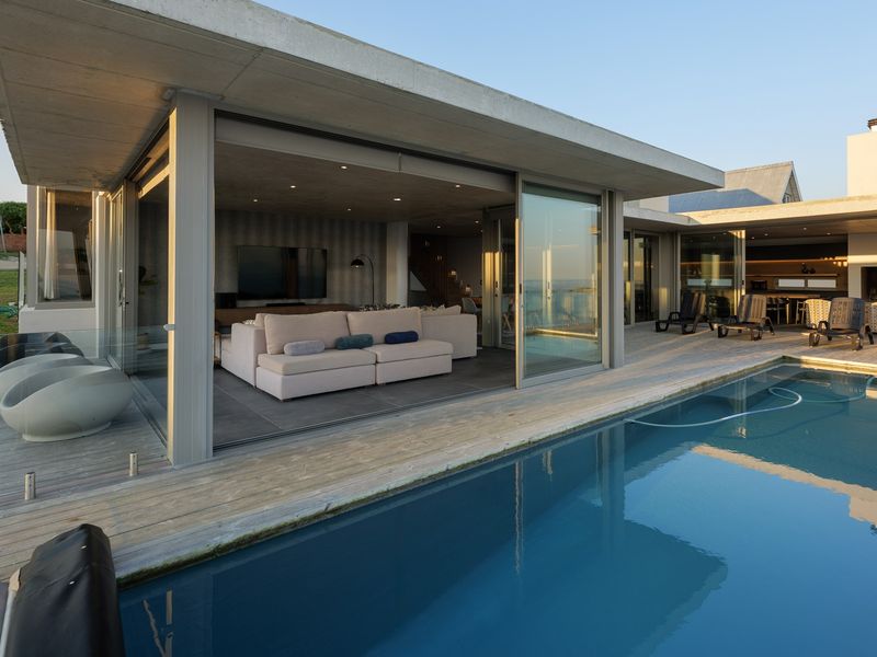 Modern outdoor pool area featuring timber decking, glass balustrades, and seamless indoor-outdoor flow through large sliding glass doors into the living space.