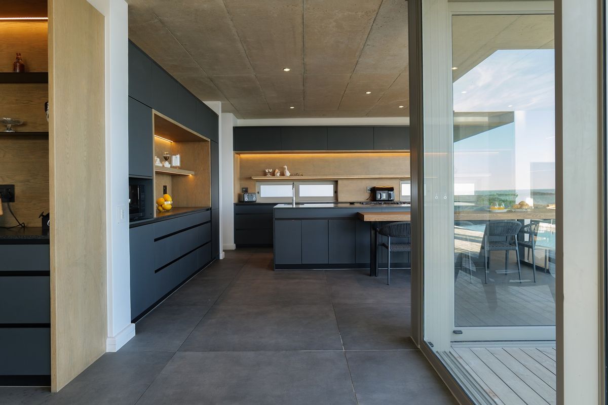 Modern, high-end kitchen featuring dark cabinetry, a central island, integrated lighting, and an exposed concrete ceiling, seamlessly connecting to an outdoor deck.
