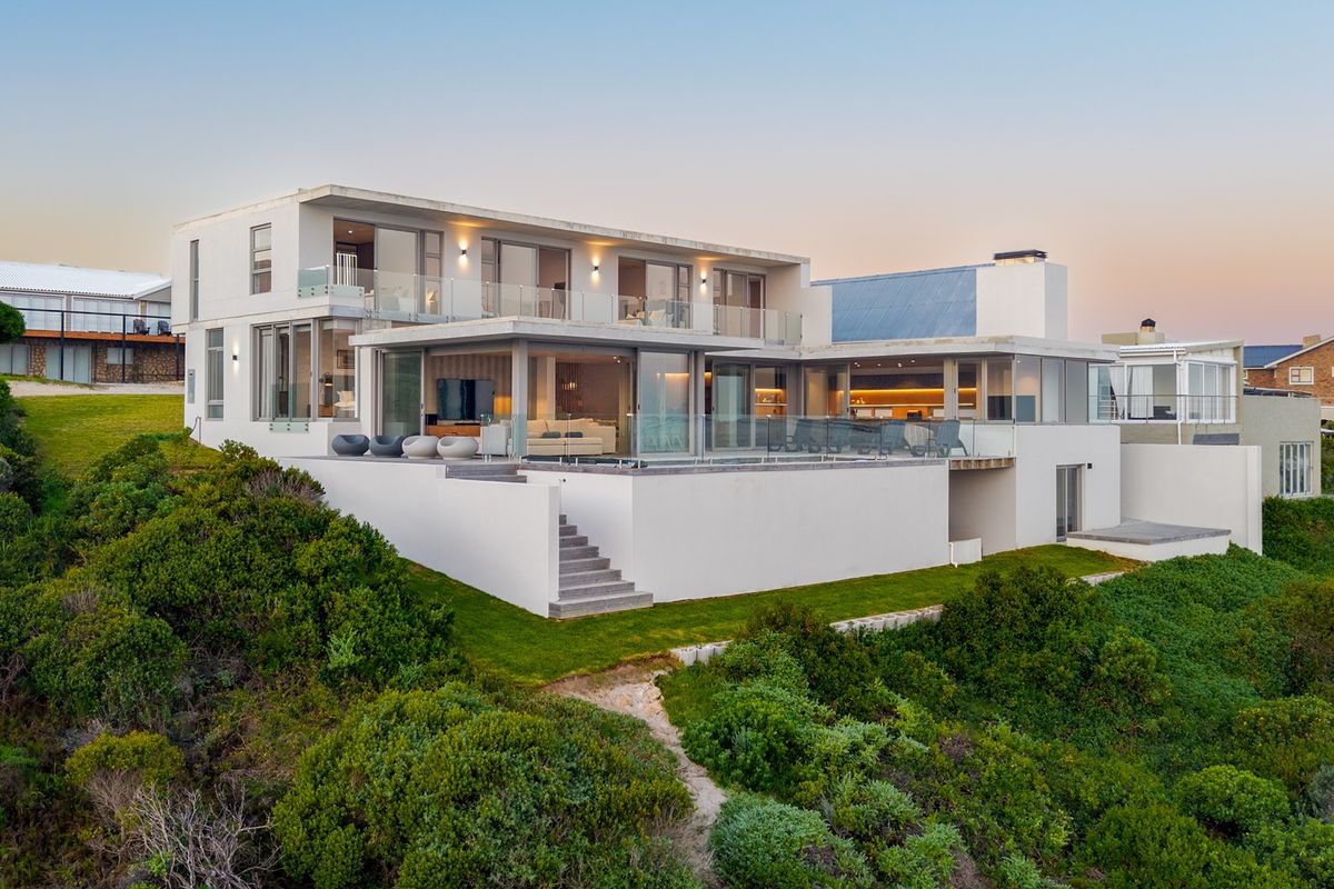 Modern multi-level coastal residence featuring expansive glass sliding doors, elevated terraces, and glass balustrades overlooking a landscaped garden.