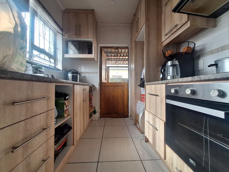 A galley-style kitchen featuring light wood-finish cabinetry, tiled flooring, and a built-in oven, leading out to a rear exterior area via a stable door.