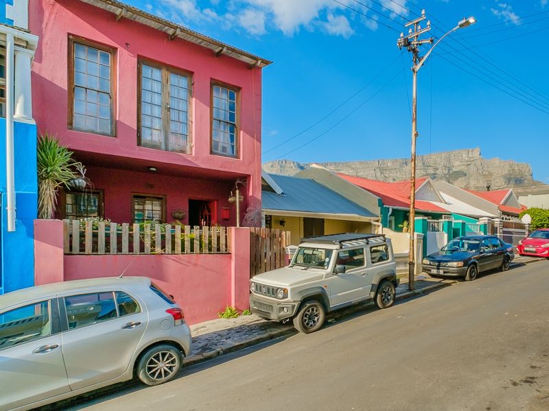 Street view of a vibrant pink two-story house with timber sash windows and a front boundary fence, set against a mountain backdrop.