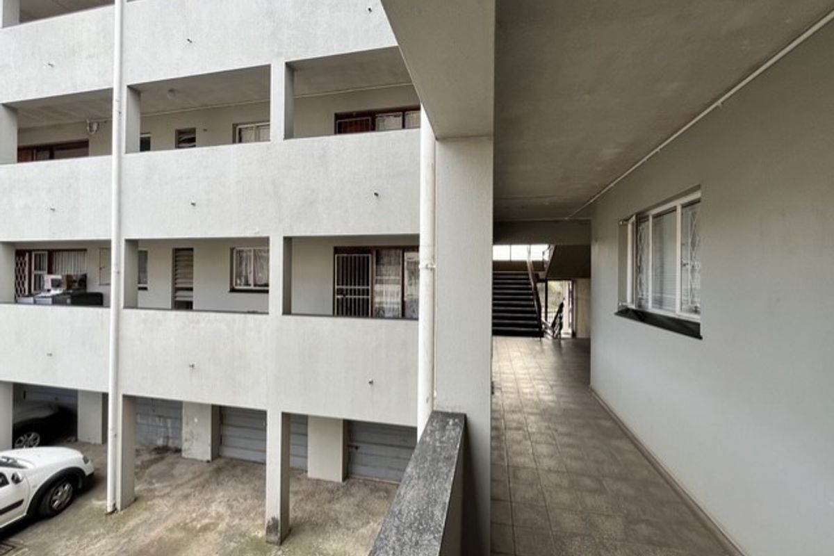 Exterior view of an apartment complex featuring a tiled walkway, concrete structure, and access to staircases and ground level parking.
